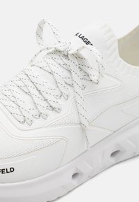 Sneaker bianca con una parte superiore in knit traspirante, dettagli con motivo triangolare, lacci piatti e una suola spessa e testurizzata con fori di ventilazione.