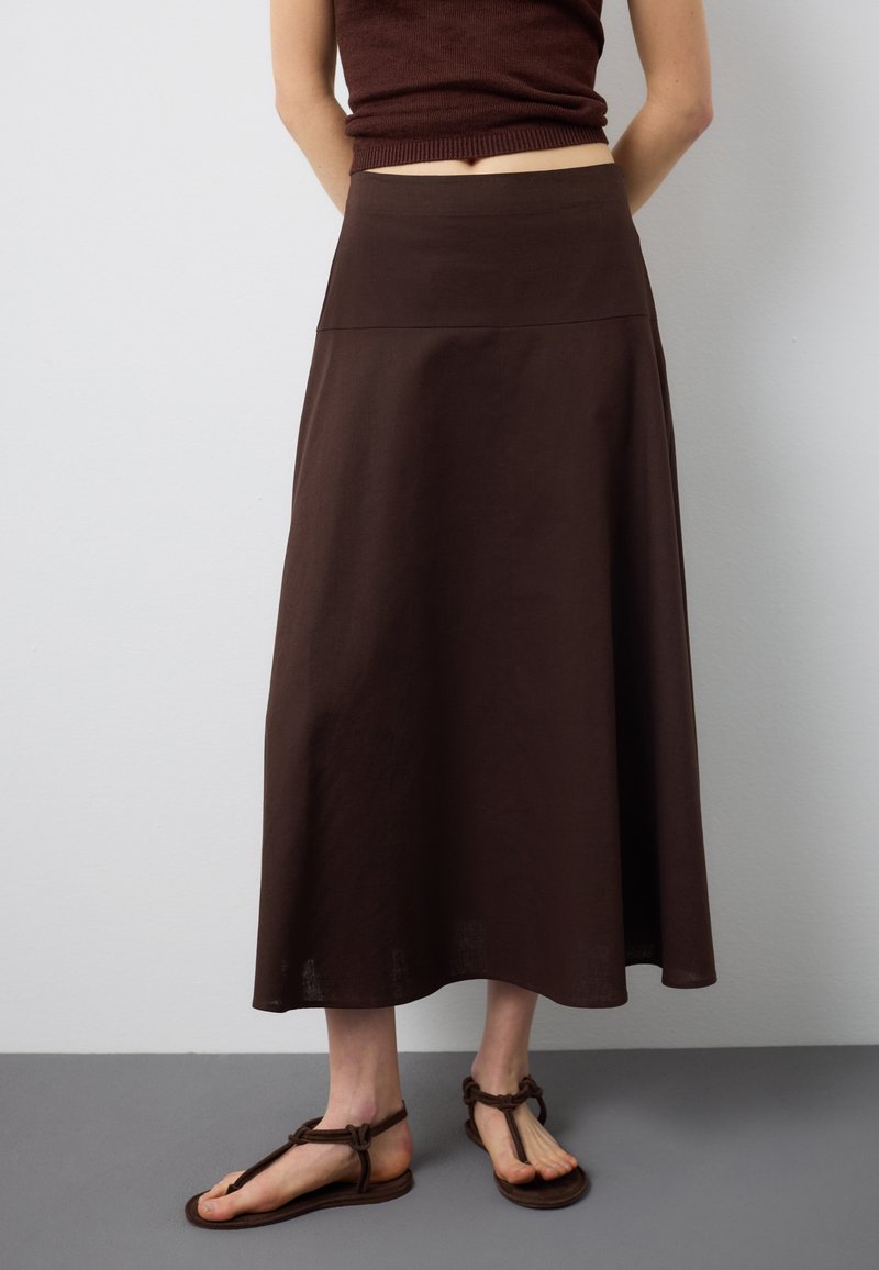 Frau trägt einen dunkelbraunen Midirock, ein ärmelloses Oberteil und passende braune flache Sandalen, steht vor einer schlichten Wand.