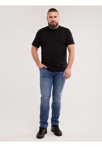 Mann mit Bart und zurückgebundenen Haaren, trägt ein schwarzes T-Shirt, blaue Jeans und schwarze Schuhe, steht mit einer Hand in der Tasche vor einem schlichten Hintergrund.