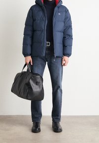 Marinblå pufferjacka med röd insida, svart tunneldoj, denimjeans och svart läderhandväska; bärande svarta stövlar.