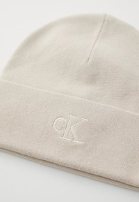 Light beige knit beanie hat with a folded cuff and embroidered "cK" logo in matching thread. Soft texture and snug fit.