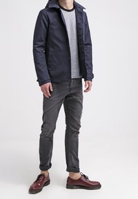 Giacca navy con colletto alto, camicia grigia testurizzata, pantaloni grigi arrotolati e scarpe borgogna con suole nere. Vestibilità casual, chiusura con bottoni.
