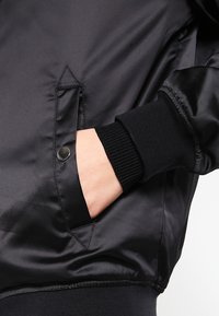 Svart satin bomberjacka med ribbad mudd och ficka med tryckknappsstängning, som visar en strukturerad yta och slät utseende.