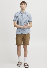 Tricou polo bleu deschis cu imprimeu cu palmieri, guler bleumarin și accente pe mâneci, asortat cu pantaloni scurți maro rulați, șosete albe și sandale maro.