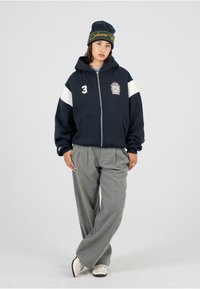 Námornícky zip-up mikina s bielymi rukávmi a vyšitým logom, v kombinácii so šedými nohavicami s širokými nohavicami a čiernym čiapkou so zlatým textom.