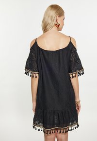 Melns off-the-shoulder kleita ar mežģīņu tekstūru, īsām piedurknēm ar pušķu apdari un volānu apakšdaļu. Parādīta no mugurpuses.