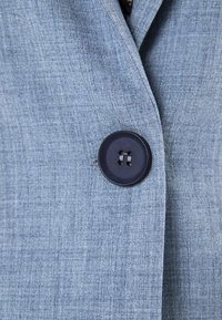 Lichtblauw jassenstof met een gestructureerde afwerking, voorzien van een grote zwarte knop met vier gaten, die eenvoudige, strakke ontwerpelementen benadrukt.