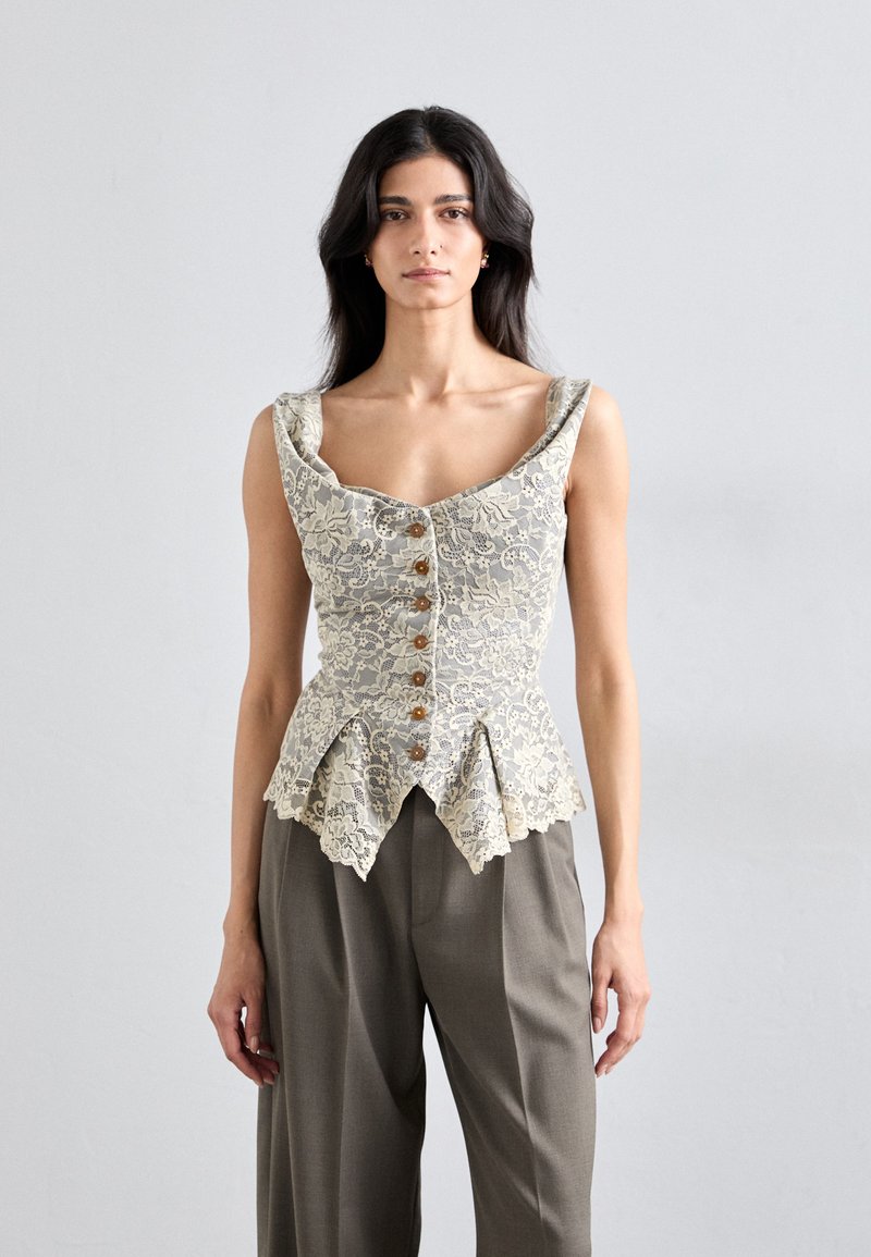 Korsetti-tyylinen pitsitoppi vaaleanbeigellä sävyllä, napituksella edessä, leveällä kaula-aukolla ja aaltoilevalla helmalla, yhdistettynä löysiin harmaisiin housuihin.