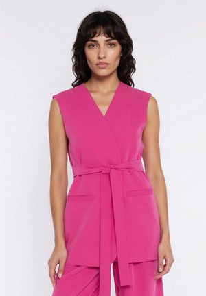 Kobieta o ciemnych, falowanych włosach nosi bezrękawnik w jaskrawym różowym kolorze z paskiem oraz pasujące spodnie na jednolitym białym tle.