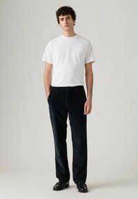 Pantaloni di velluto a coste neri con un motivo sottile, abbinati a una t-shirt bianca slim. Il modello sta in piedi, con le mani in tasca, indossando scarpe nere.