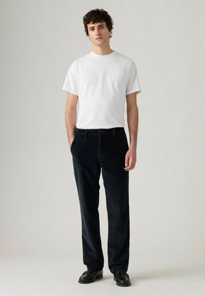 Zwarte corduroy broek met een subtiele patroon, gecombineerd met een getailleerd wit T-shirt. Model staat met de handen in de zakken en draagt zwarte schoenen.