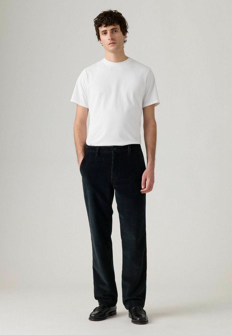 Pantaloni di velluto a coste neri con un motivo sottile, abbinati a una t-shirt bianca slim. Il modello sta in piedi, con le mani in tasca, indossando scarpe nere.
