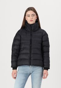 Swarte puffjakke med høy krage, frontglidelås og sidelommer. Har quiltede seksjoner og ribbede mansjetter. Jevn, matt tekstur.