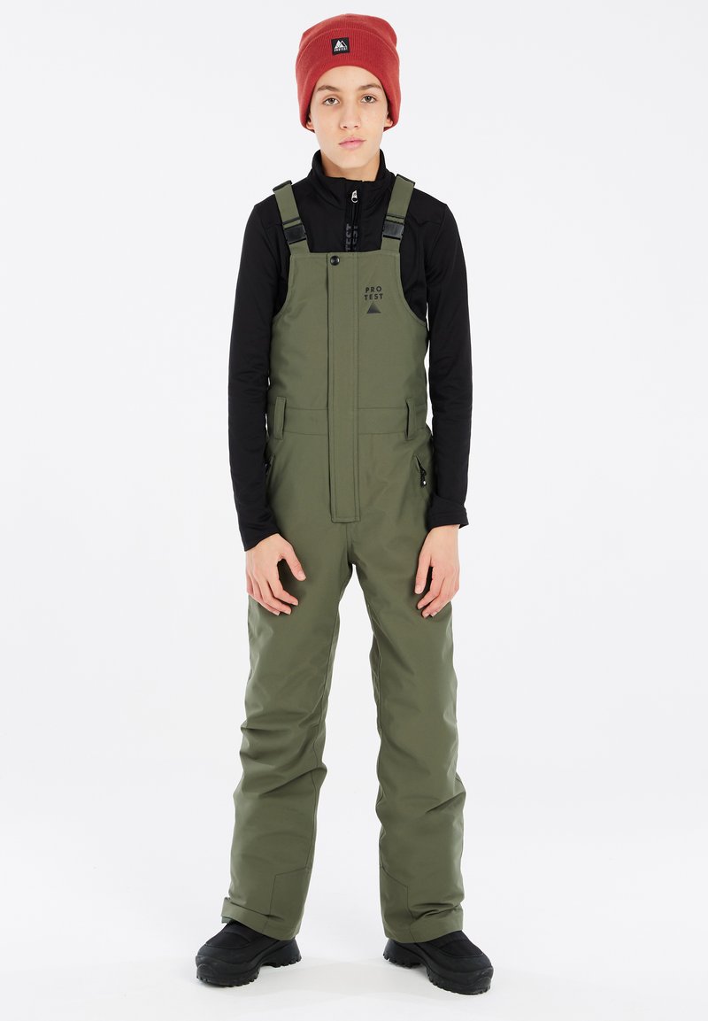 Salopette verde con spalline regolabili, tasche laterali e chiusura con zip, indossata sopra una maglietta nera a maniche lunghe e abbinata a un berretto rosso.