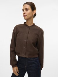 Femme portant une veste bomber marron à boutons avec des poignets et un col côtelés, associée à un jean bleu foncé, se tenant devant un fond blanc.