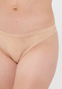Taille en bovenbeen van een persoon in lichtbeige katoenen ondergoed, waarbij de natuurlijke huidtextuur en kleine moedervlekken zichtbaar zijn.