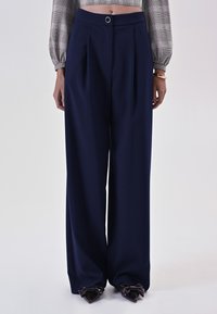 Pantaloni a gamba larga color navy realizzati in tessuto testurizzato; presentano una parte frontale plissettata e un dettaglio a bottone circolare in vita. Accentuati da scarpe a forma di fiocco.