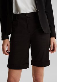 Nainen pukeutuneena mustiin räätälöityihin polvimittaisiin shortseihin, joissa on käännetyt lahkeet, yhdistettynä mustaan bleiseriin ja valkoiseen puseroon.