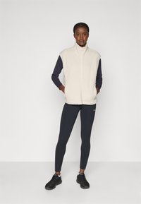 Fleece vest in crèmekleur met een hoge kraag, ritssluiting aan de voorkant en zijzakken, gecombineerd met donkere leggings en zwarte sportschoenen.
