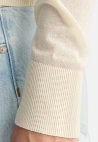 Polsino in maglia crema con texture a coste e un sottile dettaglio del logo. Abbinato a jeans denim azzurri, che mostrano una vestibilità rilassata e un tessuto morbido.