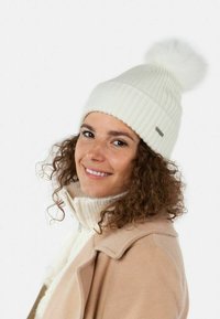 Beanie bianco a coste con pompon in pelliccia sulla cima. Materiale caldo, design aderente. Dettaglio di etichetta argento visibile sul lato.
