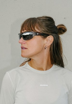 Femme portant des lunettes de soleil noires enveloppantes et un t-shirt blanc Salomon, représentée de profil devant un mur uni.