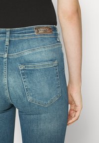 Vaqueros de denim azul con una cintura alta, que presentan un parche de cuero marrón en el logo y bolsillos traseros clásicos con un discreto detalle de costura.