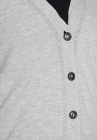 Šedý kardigan vyrobený z jemné látky, s výstřihem do V, pěti černými knoflíky a žebrovanou texturou po okrajích.