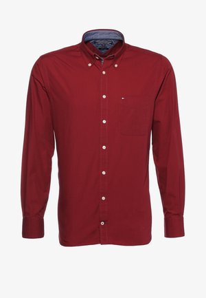 Ein Mann, der läuft, trägt ein rotes Hemd mit Knöpfen, graue Jeans mit Umschlägen, einen schwarzen Gürtel, graue Turnschuhe und eine Armbanduhr.
