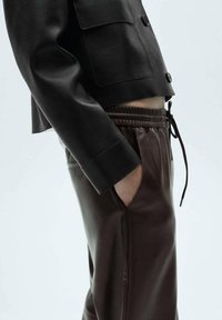 Veste courte en cuir noir avec deux poches poitrine, associée à un pantalon de survêtement marron avec une ceinture élastique et des poches latérales.