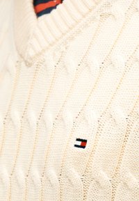 Πουλόβερ σε κρεμ απόχρωση με πλεχτό σχέδιο καλωδίου, με λαιμόκοψη σε σχήμα V και μικρό κεντημένο λογότυπο σε κόκκινο, λευκό και μπλε.