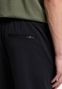 Schwarze Sporthose mit einer Gesäßtasche und einem Etikett. Das Material wirkt weich, mit einem elastischen Bund und einem schlanken Design.