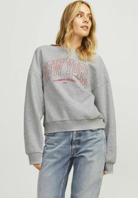 Felpa grigia con scollo rotondo, vestibilità oversize e scritta rossa "NEW YORK". Presenta polsini e orlo a coste; indossata con jeans azzurri.