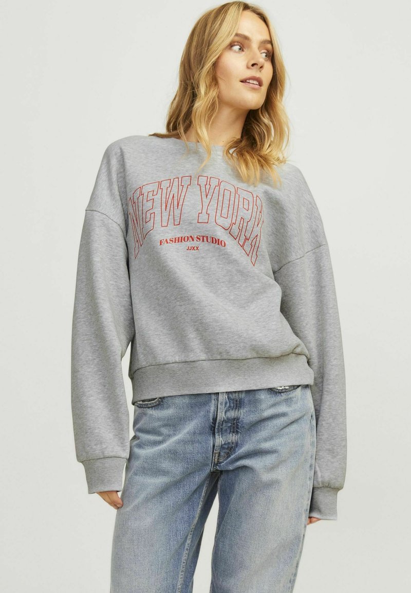 Felpa grigia con scollo rotondo, vestibilità oversize e scritta rossa "NEW YORK". Presenta polsini e orlo a coste; indossata con jeans azzurri.