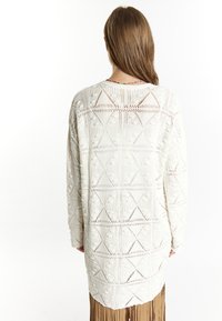 Långärmad, vit stickad tröja med geometriska mönster och texturerade detaljer. Har en öppen design och en avslappnad passform.