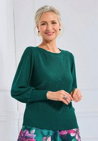 Grüner Strickpullover mit strukturiertem Finish, geripptem Kragen und Bündchen sowie glitzernden Details. Kombiniert mit einem blumengemusterten Rock.