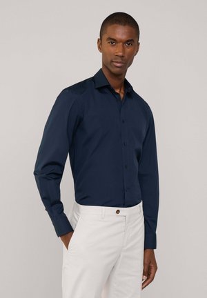 Camisa de manga larga azul marino de tela suave, con cierre de botones en el frente y cuello puntiagudo. Combinada con pantalones blancos.