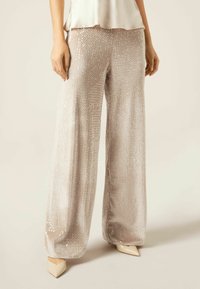 Pantalony s širokými nohavicemi v světlé béžové barvě, s texturovaným povrchem pokrytým malými kulatými flitry. Spárovány s béžovými lodičkami s špičatou špičkou.