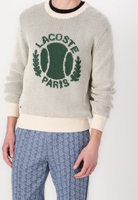 Stickad tröja i crème med grön "LACOSTE PARIS"-logotyp. Har ribbade mudd och nederkant. Kombineras med byxor i blått mönster.