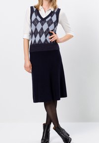 Donkerblauwe gebreide vest met ruitpatroon over een witte blouse met kraag, gecombineerd met een donkerblauwe knielange rok en zwarte hoge enkellaarsjes.