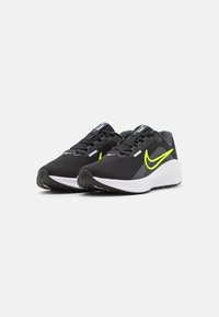Sorte atletiske sko med en struktureret mesh-overdel, lys gul swoosh, snørebånd og en hvid mellemsål, der har en mønstret sort ydersål.