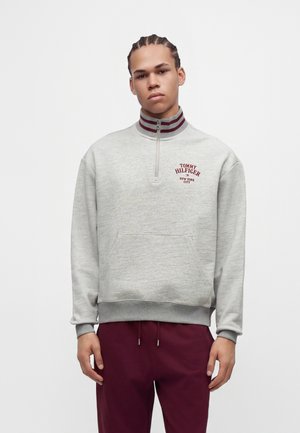 Млад мъж с плитки, облечен в сива блуза с цип до средата и лого Tommy Hilfiger и бордо панталони, стоящ на бял фон.