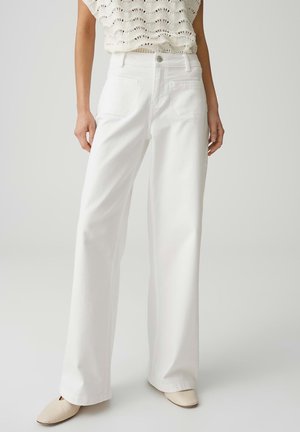 Frau trägt weiße, hoch taillierte, weite Hose, gestricktes ärmelloses Oberteil und hellbeige spitze Schuhe, steht vor einem schlichten, hellen Hintergrund.