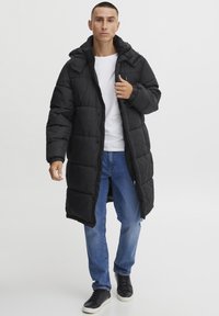 Svart boblejakke med hette, med glidelås og knappelukking, quiltet tekstur og sidelommer, båret over en hvit skjorte og blå jeans.