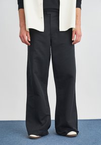 Pantaloni neri a gamba larga con texture liscia, caratterizzati da dettagli di cucitura laterale e una silhouette rilassata, abbinati a sneaker bianche.