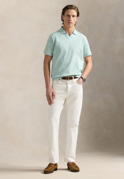 Φωτεινό γαλάζιο πόλο πουκάμισο, λευκό τζιν, καφέ παπούτσια τύπου slip-on. Το μοντέλο στέκεται με το ένα χέρι στην τσέπη. Απλός σχεδιασμός, απαλή υφή και ουδέτερα χρώματα.