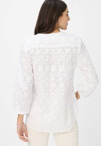 Weiße bestickte Bluse mit floralen Mustern und 3/4 Ärmeln. Leichter Stoff mit einer sanften Textur und gekurvtem Saum.