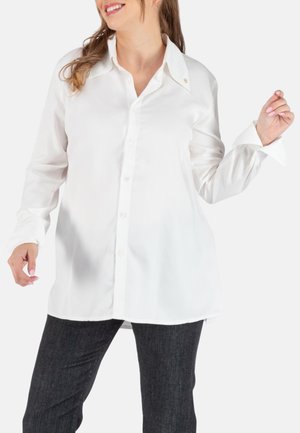Weiße Bluse mit Knopfleiste und Kragen, lockerer Schnitt, lange Ärmel und einem dezenten Logodetail in der Nähe des Kragens. Kombiniert mit dunklen strukturierten Hosen.