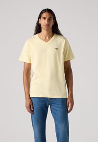 Tricou galben cu decolteu în V, din bumbac, cu mâneci scurte, având un mic patch cu logo-ul roșu Levi's lângă gât. Purtați cu blugi bleumarin.