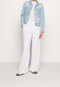 Veste en denim bleu clair avec des accents usés, portée sur un T-shirt gris clair. Un pantalon large blanc et des sandales noires à enfiler complètent le look.